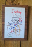 A framed vintage-style embroidered art piece featuring a cartoon dog standing on a fence with the word 'Friday' in red script at the top, displayed on a wooden surface.