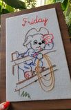 A rectangular embroidered art panel mounted on a wooden frame, featuring a cartoon dog standing on a fence with the word "Friday" stitched in red at the top. The embroidery uses multiple thread colors including blue, red, yellow, green, and brown on a light fabric background, photographed with a hand visible at the bottom left and greenery in the background.
