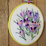 A circular embroidery hoop containing a floral embroidered design in purple, pink, and green on cream fabric, photographed against a rustic wooden background.