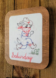 A square wooden framed art with rounded corners featuring a white center panel with a hand stitched cowboy dog wearing a hat, bandana, and boots, with the word 'Saturday' stitched in red at the bottom. The plaque is photographed on a rustic wooden surface.