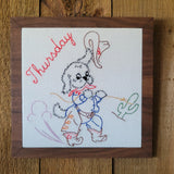 A square framed piece of embroidered art featuring a cartoon dog dressed in cowboy clothing with the word 'Thursday' in red script. The embroidery is on a white background and is housed in a dark brown wood frame, set against a rustic wooden wall.