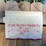 An off-white fabric wall hanging with pink embroidered text reading 'I Am Too Pretty For This Shit' and a pink floral design, hanging from a wooden fence outdoors.