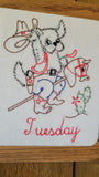 A square white embroidered frame featuring a cartoon dog riding a rocking horse, with a cowboy hat, boots, cactus, and the word "Tuesday" stitched in red at the bottom, shown on a wooden surface.