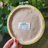 A close-up of a vintage needlework hoop containing light beige wool felt, held by a hand with red nail polish. The center features a white paper label that reads 'Vintage Needlework Repaired / Rebuilt by: EMBER 6 THREAD'.