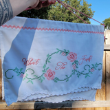 A white embroidered wall hanging outdoors on a wooden fence. The fabric features pink embroidered roses, green leaves, and the words 'What The Fuck?' in pink script.