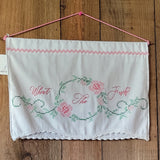 A white fabric wall hanging or decorative panel with pink embroidered script reading 'What The Fuck?' inside a floral wreath design, with pink and green embroidery, a pink hanging cord, and a lace-trimmed bottom edge, displayed against a wooden plank background.