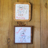 Two square wooden framed art with rounded corners displayed on a wooden surface. The top one has a light wood frame and a white center with a hand-drawn cartoon dog and the word 'Wednesday' in red script.