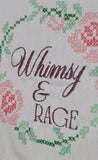 Close-up of a white fabric item featuring embroidered floral patterns in green and pink, with the words 'Whimsy & RAGE' stitched in dark grey cursive and serif lettering.