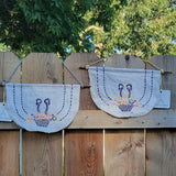 Two white fabric banners with embroidered baskets and bows, hanging on a wooden fence outdoors.