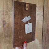 A dark brown wooden board with a small wooden keyhole-shaped piece at the top, held against a wood-paneled background by a hand with red nail polish.