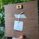 A wooden picture frame backplate with a natural wood keyhole cutout, silver tape, and a white tag reading 'EMBER & THREAD', held by a hand with red nail polish in front of green plants.