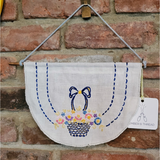 A handmade embroidered textile wall hanging featuring a basket of colorful flowers and blue bows, suspended from a wooden rod against a red brick wall. A white paper tag on the right reads 'EMBER & THREAD'.