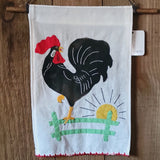 A white fabric wall hanging featuring a stylized black rooster with a red comb, standing above a green fence and yellow sun, hanging from a wooden dowel on a wood plank background.