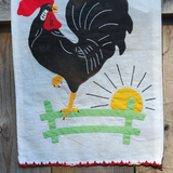 A white fabric wall hanging featuring a stylized black rooster with a red comb, yellow beak and legs, a green fence, and a yellow sun on a rustic wooden background.