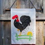 A white fabric wall hanging on a wooden dowel on a rustic wooden fence. The flag features a stylized black rooster with a bright red comb, a yellow beak and legs, a green fence, and a yellow sun with black rays.
