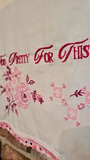 A close-up of an off-white fabric textile with pink embroidered script reading 'Too Pretty For This' and pink floral motifs, finished with a pink lace trim along the edge.