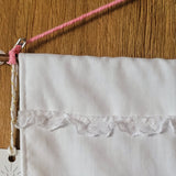A close-up of a white fabric wall hanging with a decorative sparkly white lace trim across the middle, suspended from a wooden rod with a pink cord against a wood-grain background.