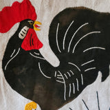A close-up of a wall hanging featuring a stylized rooster design in black with a bright red comb and wattle, white stitched feather lines, and small yellow accents on a white background.