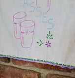 A close-up of a white embroidered fabric featuring pink outlined tumblers, blue stitched text, green leaves, and purple flowers, with a decorative purple and green scalloped border along the bottom edge.