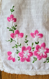 A close-up of a white embroidered fabric, featuring pink flowers and green leaves stitched along the border.