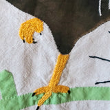 Close-up of a handmade embroidered textile featuring a stylized bird with a yellow-orange beak and leg, black details, a brown upper section, and light green and off-white lower sections.