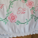 Close-up of a white embroidered textile with pink floral motifs, green vine stitching, and partial pink script embroidery, finished with a white lace trim along the bottom edge.
