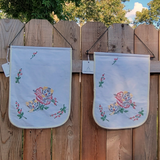 Two white fabric flags with colorful embroidered flowers and cats, hanging from a wooden fence outdoors.