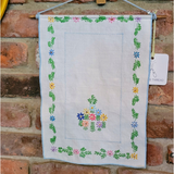 A white wall hanging with embroidered floral borders and a central floral motif, suspended against a rustic brick wall.