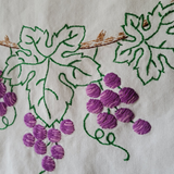 Close-up of embroidered grapevine and grape clusters on a white fabric surface, with green leaf outlines, brown vine details, and purple stitched grapes.