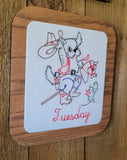 A square wooden plaque with rounded corners featuring a white center panel with a hand-embroidered cartoon dog wearing cowboy boots and a hat, riding a small horse, with the word 'Tuesday' embroidered in red at the bottom. The plaque is photographed on a rustic wooden surface.