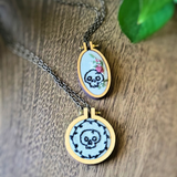 Two oval pendant necklaces with skull embroidery in wooden hoop frames, displayed on a rustic wood surface with a green leaf in the background.