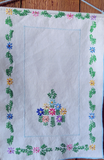 A square white fabric featuring colorful embroidered flowers and green leaves around a border, with a central floral motif and a light blue stitched inner rectangle, laid flat on a wooden surface.