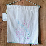 A white fabric vintage wall hanging with embroidered pink glasses and the word 'Glasses' on it, hanging on a wooden background