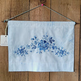 A square light blue dyed fabric wall hanging with a blue floral toile-style print across the center, suspended by a cord against a wooden plank background.