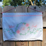 A white fabric wall hanging with pink and green embroidered details, hanging from a wooden fence outdoors. The banner features the text 'What The Fuck?' in pink script, surrounded by a floral vine design.