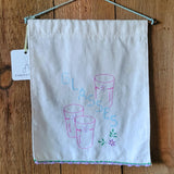 A white fabric wall hanging with embroidered pink and blue glassware and the word 'GLASSES,' shown hanging on a wooden wall with a small product tag attached.