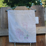 A white cotton tea towel with embroidered glass cups and flowers, repurposed into a wall hanging