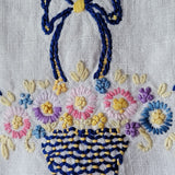 Close-up of a hand-embroidered basket of flowers on a white linen fabric background. The embroidery features navy blue, yellow, pink, light blue, purple, and cream threads.
