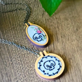 Two handmade oval necklaces with wooden hoop pendants featuring embroidered black skulls on a grey fabric background, resting on a wooden surface.