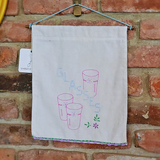 A white fabric wall hanging panel with embroidered pink and blue glassware and the word "Glasses," hung against a red brick wall.