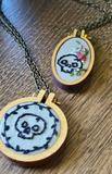 Two oval wooden hoop necklaces with embroidered skull and floral designs on a wooden table. Both necklaces feature black skulls, both hanging from dark metal chains.