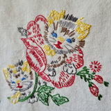 Close-up of embroidered floral and feline artwork on a light fabric background, featuring a large red flower, smaller red buds, green leaves, and two stylized cat faces in yellow, gray, blue, red, brown, and white stitching.