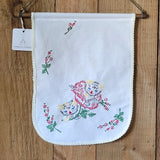 A white cotton wall hanging with embroidered kittens and floral motifs, hanging from a wooden dowel on a rustic wooden background.