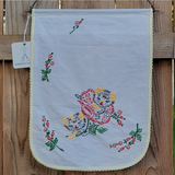 A square white fabric with scalloped edges and colorful floral embroidery, hanging on a wooden fence.