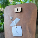 A close-up of a wooden square peg with a central hole, mounted on a wooden board. A small white tag with the brand name 'EMBER & THREAD' is on the back