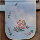 A vintage embroidered wall hanging with a scalloped cream border, featuring colorful floral and kitten embroidery on a white background, hanging on a wooden surface.