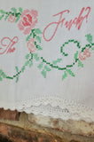 Close-up of a white embroidered textile with pink roses, green vine stitching, and pink script lettering, finished with a white lace trim along the hem.