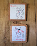 Two square wooden framed prints displayed on a wooden surface. The top card is labeled 'Monday' and shows a cowboy dog playing a guitar.