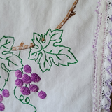 Close-up of a white embroidered textile featuring green grape leaves and stems with purple embroidered grapes, alongside a lavender lace trim.