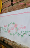 A white embroidered wall hanging with pink script text reading 'What The Fuck?' inside a floral wreath design with green leaves, hanging against a brick wall.